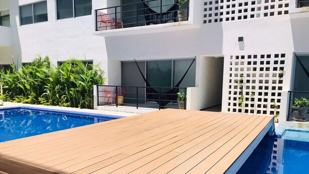 Uma área de piscina moderna ao ar livre com deck de madeira, cercada por vegetação exuberante e um edifício branco com sacadas.