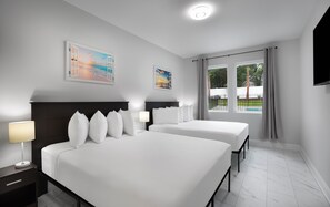Quarto de hotel com duas camas, uma mesa de cabeceira, uma luminária e um quadro de uma praia.