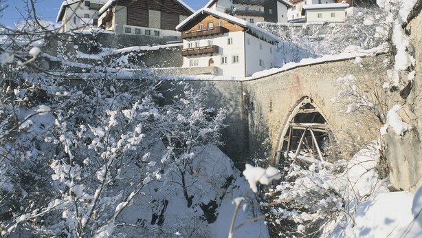 A snow-covered mountain village with traditional wooden houses, a stone bridge, and a clear blue sky. - Apartment Huber by Interhome (Grins)