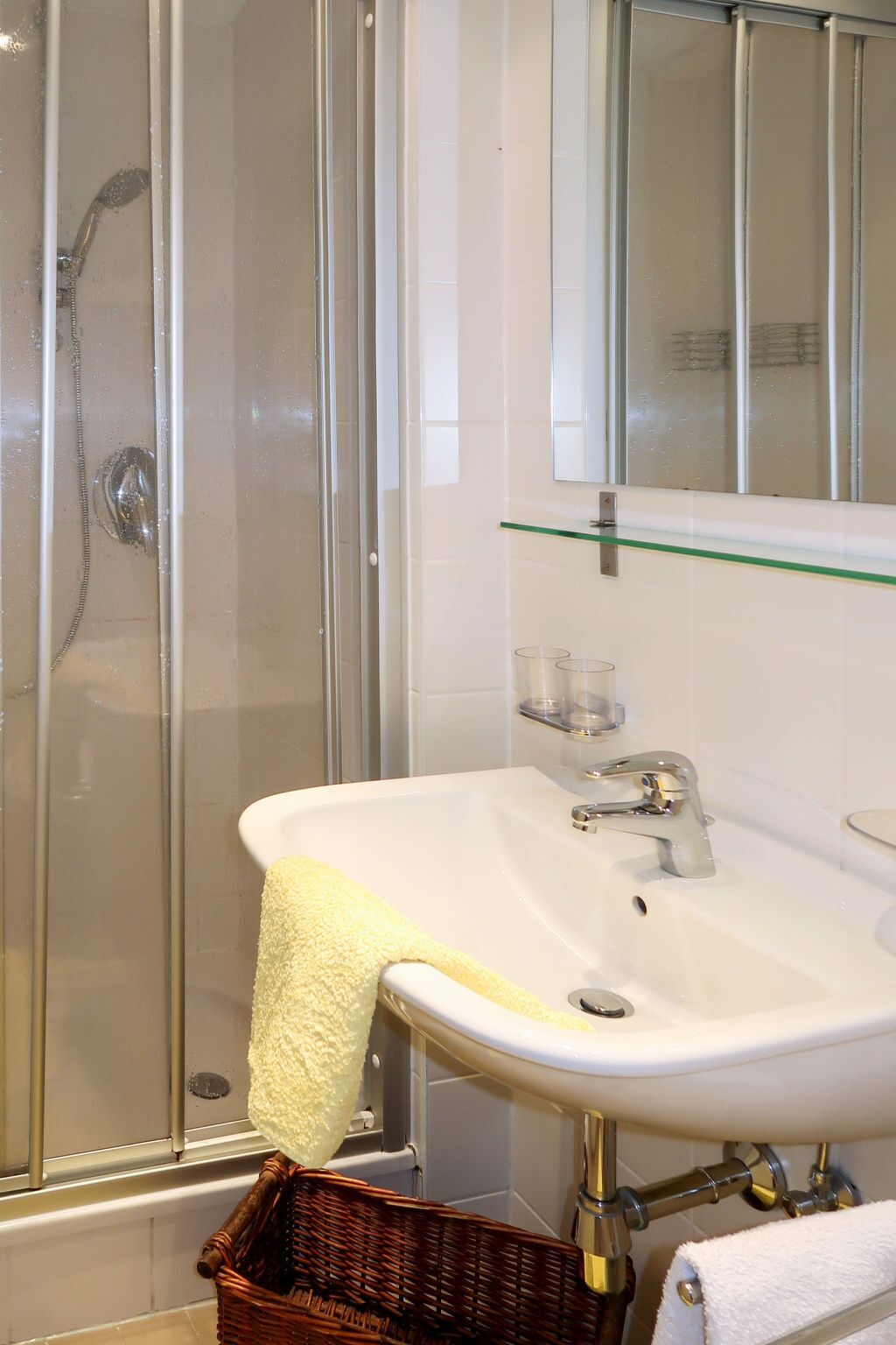 A bathroom with a glass shower enclosure, a white sink with a gold faucet, and a glass shelf above the sink.