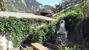A rustic outdoor seating area with wooden benches and a stone table, surrounded by lush greenery and a mountain backdrop. - Martina by Interhome (Pfunds-Samnaun)