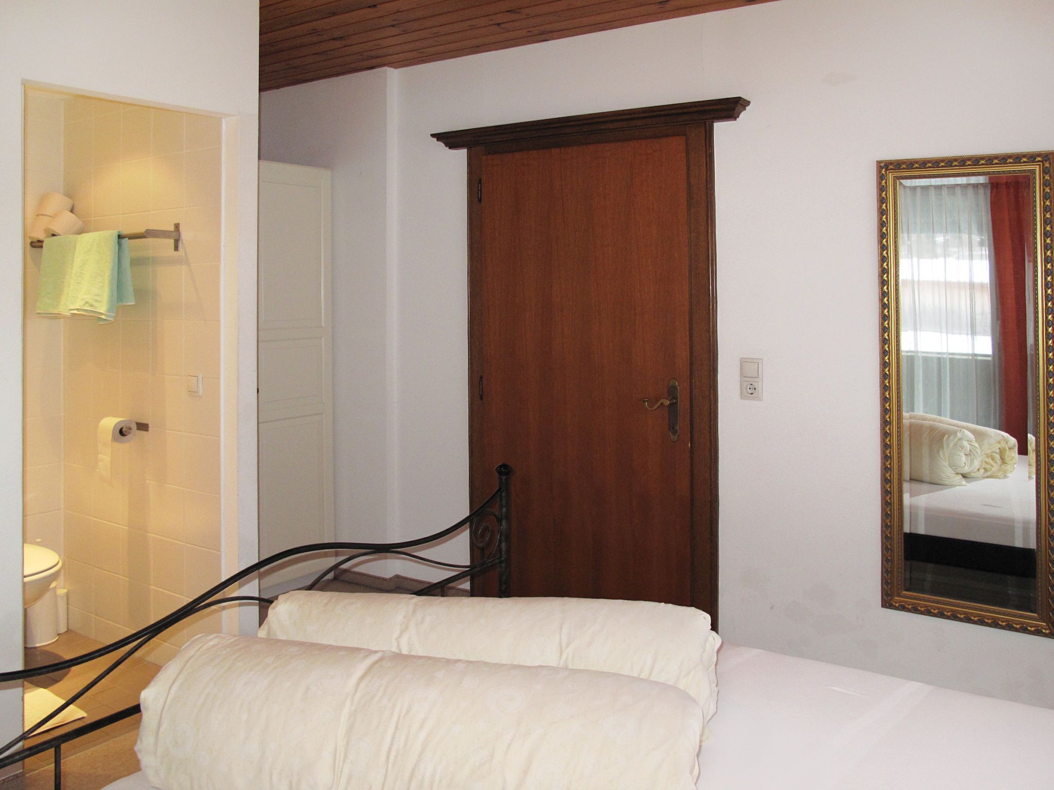 A bedroom with a wooden door, a mirror with a decorative frame, and a bed with white bedding.