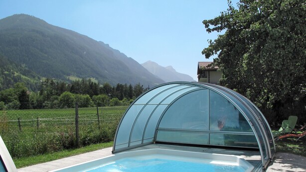 A swimming pool with a glass-enclosed hot tub, surrounded by a lush green landscape and mountains in the background.