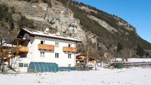 A two-story chalet with wooden balconies, a snow-covered ground, and a mountainous backdrop.