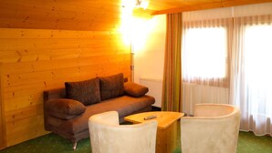 A cozy room with a wooden wall, a sofa, two armchairs, a small wooden table, and a window with curtains.