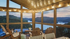 A wooden deck with Adirondack chairs and a table set for dining, overlooking a lake and forested hills.