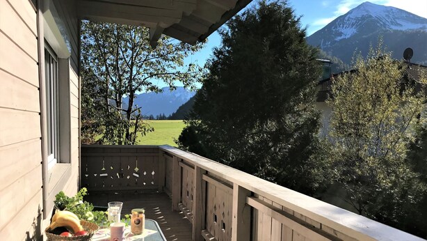 Un balcón con una mesa lista para dos, con vistas a una montaña y un campo verde.