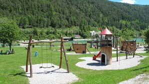 A playground with swings, slides, and climbing structures, surrounded by a grassy area and a forested hill. - Apartment Rofan by Interhome (Achensee)