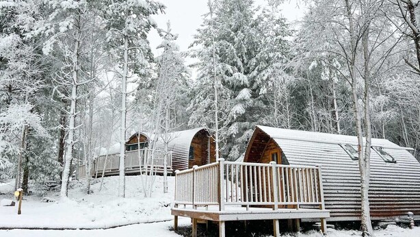 A row of log cabins with wooden decks and railings, surrounded by snow-covered trees.