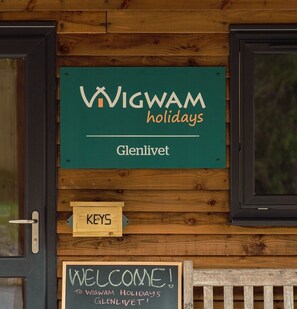 Eine Holzwand mit einem Schild, auf dem 'WIGWAM holidays Glenlivet' steht, und einem Kreideschild mit der Aufschrift 'WILLKOMMEN! BEI WIGWAM HOLIDAYS GLENLIVET!'.