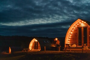 Dos cabañas de madera con interiores iluminados, una mesa de picnic y un banco.