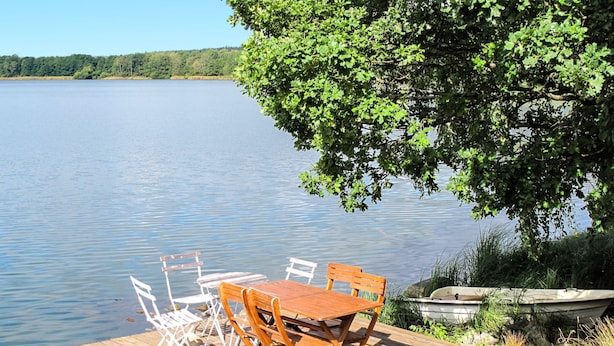 Une jetée en bois avec une table et des chaises, donnant sur un lac paisible, entourée de verdure et avec un bateau à proximité.