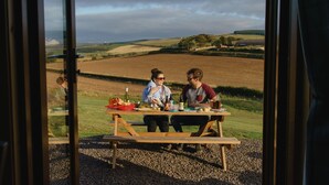Una mesa de picnic de madera con comida y bebidas, con un paisaje rural de campos y colinas al fondo.