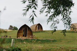 Dos cabañas de madera con forma de cúpula, cada una con un banco en frente, ubicadas en un campo cubierto de césped y con árboles al fondo.