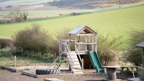 Una estructura de madera con tobogán y red de escalada, rodeada de grava y árboles, frente a un paisaje rural.