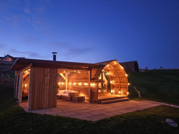 Una cabaña de madera acogedora con techo curvo, chimenea y zona de estar con cojines.