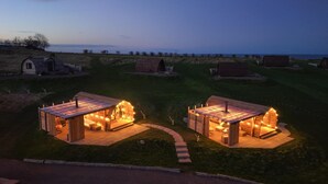 Due cabine in legno con portici, ciascuna dotata di un piccolo braciere e luci a stringa, immerse in un'area erbosa con vista sul mare.