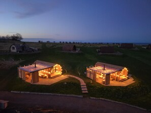 Twee houten vakantiehuisjes met aanbouwen, elk met een kleine vuurkuil en lichtjes, gelegen in een grasrijk gebied met uitzicht op zee.