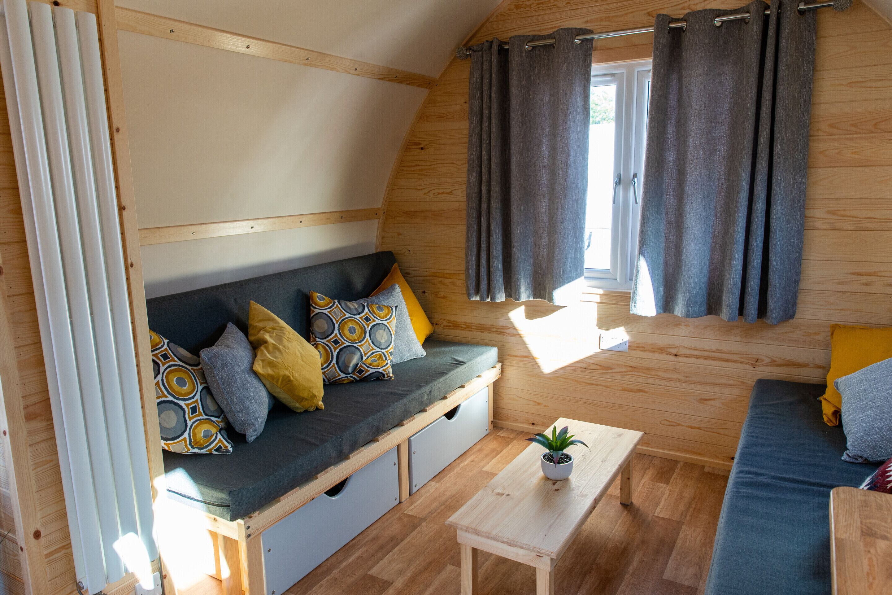 A cozy cabin interior with a wooden interior, a sofa with cushions, a small table with a plant, and a window with curtains.