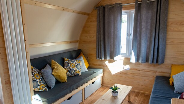 Een gezellig chaletinterieur met houten details, een bank met kussens, een kleine tafel met een plant en een raam met gordijnen.