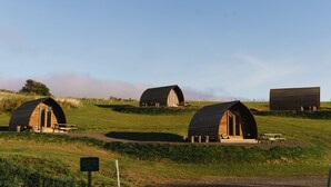 Een rij houten hutten met picknicktafels, gelegen in een grasveld onder een heldere lucht.