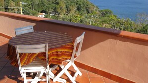Una terraza con mesa y sillas, con vistas a un paisaje costero con colinas y el mar.