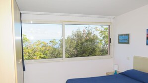 Un dormitorio con cama, ventana con vistas a árboles y al mar, y una mesita de noche con lámpara.