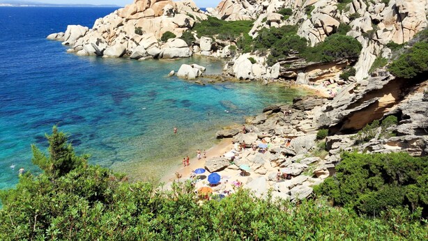 A rocky coastline with clear blue waters and a beach area with umbrellas and people.