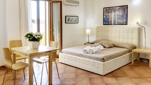 Un dormitorio con cama, mesita de noche, lámpara, silla, una mesa con un florero y una ventana con cortinas.