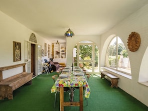 Un espace repas avec une longue table, des chaises et un tapis vert. Il y a un banc le long du mur, une porte et une fenêtre donnant sur un paysage verdoyant.