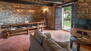 Interior rústico de piedra con pared de piedra, vigas de madera, un televisor grande y una zona de comedor con bancos y una mesa.