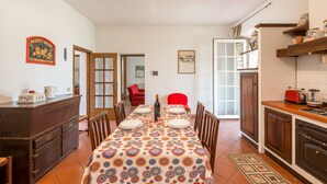 A dining area with a patterned tablecloth, wooden chairs, a red chair, a kitchen with a stove, and a cabinet with kitchenware.