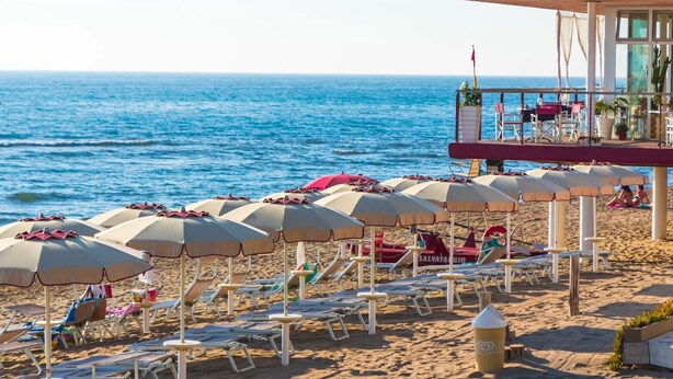 Une vue sur la plage, avec des rangées de parasols et de chaises longues, un poste de surveillance des sauveteurs, et l’océan en arrière-plan.