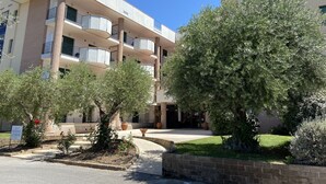 Un immeuble résidentiel à plusieurs étages, doté de balcons, d’un jardin aménagé et surplombant un ciel d’un bleu éclatant.