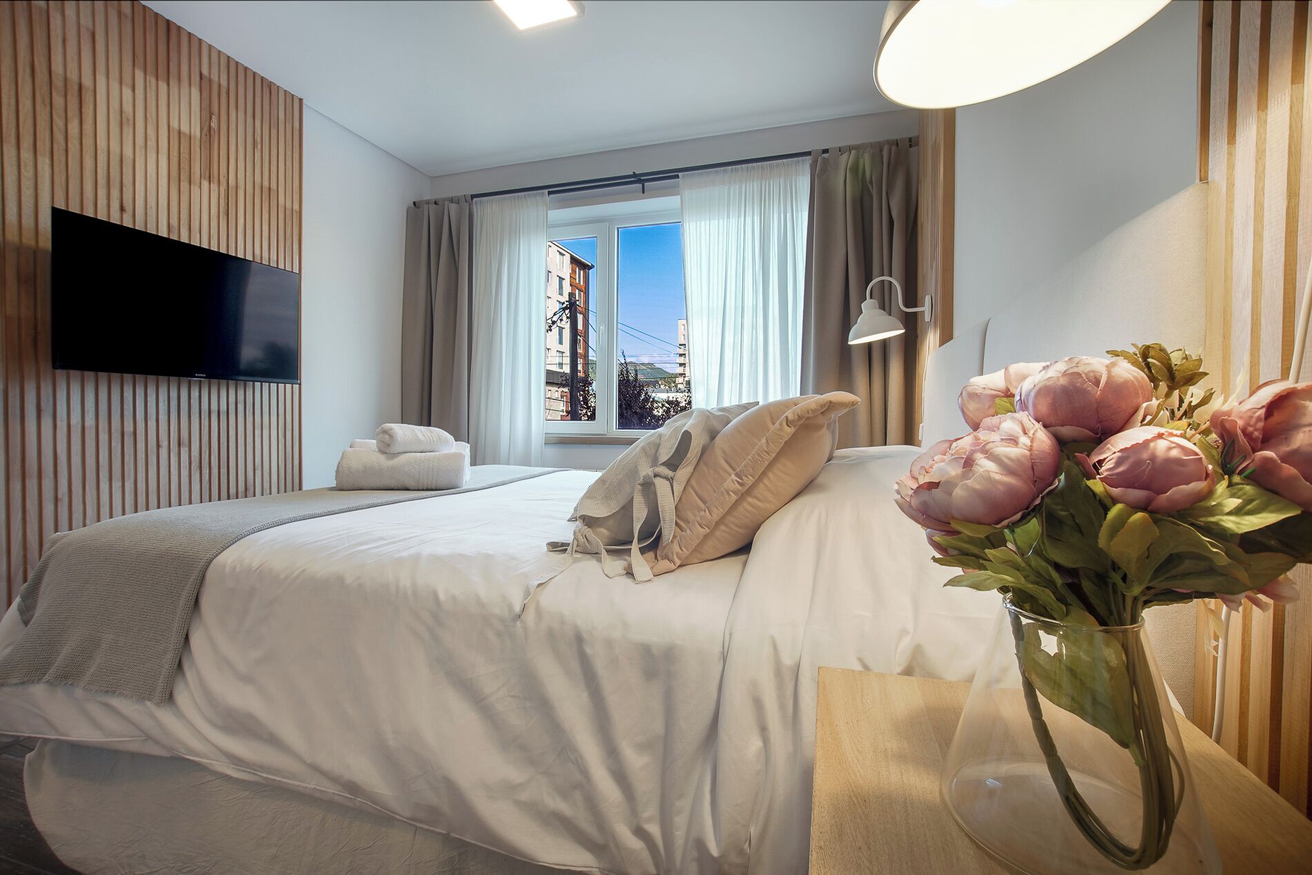 Een moderne hotelkamer met een bed, een aan de muur bevestigde televisie, een raam met gordijnen en een nachtkastje met een bloemenvaas.