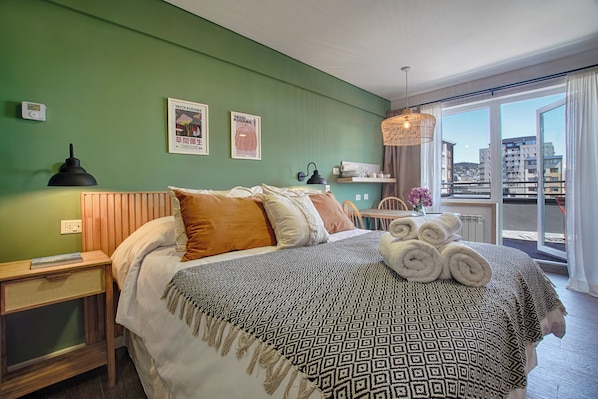 A modern bedroom with a green wall, a wooden headboard, a patterned bedspread, and a view of the city through large windows. - Edificio Montaña (San Carlos de Bariloche)