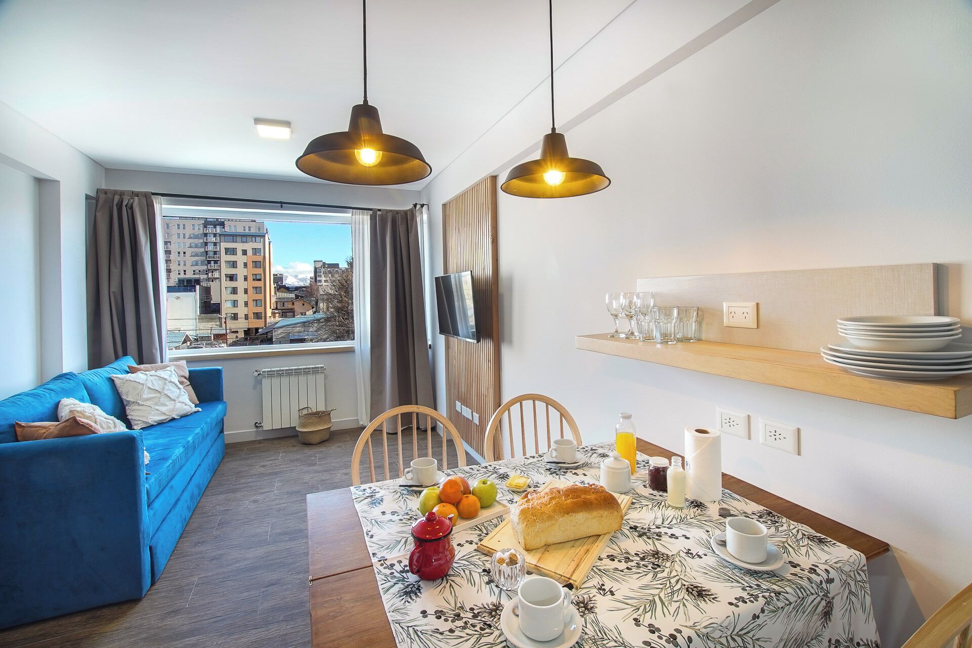 A modern living room with a blue sofa, a dining table set with fruits, a loaf of bread, and a TV mounted on the wall.