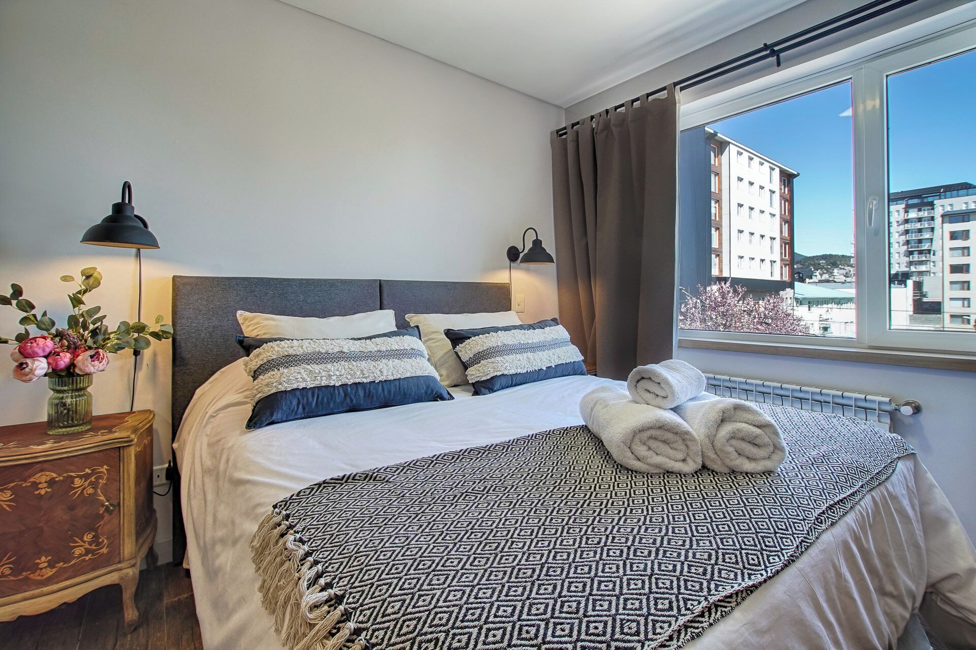 A neatly made bed with a patterned blanket, two bedside lamps, a vase with flowers, and a view of buildings through a window.
