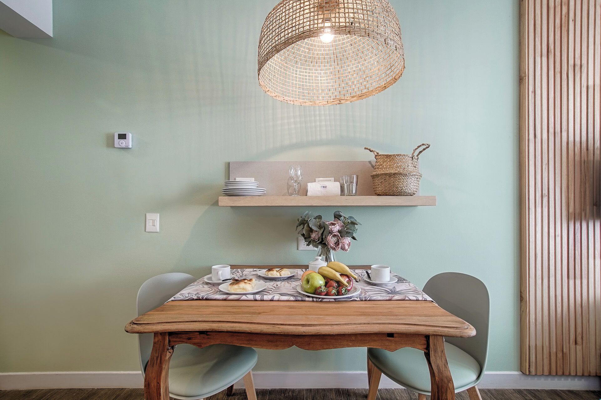 Uma sala de jantar com uma mesa de madeira, duas cadeiras, um candeeiro tecido pendurado e uma prateleira na parede com pratos, copos e uma cesta de vime.