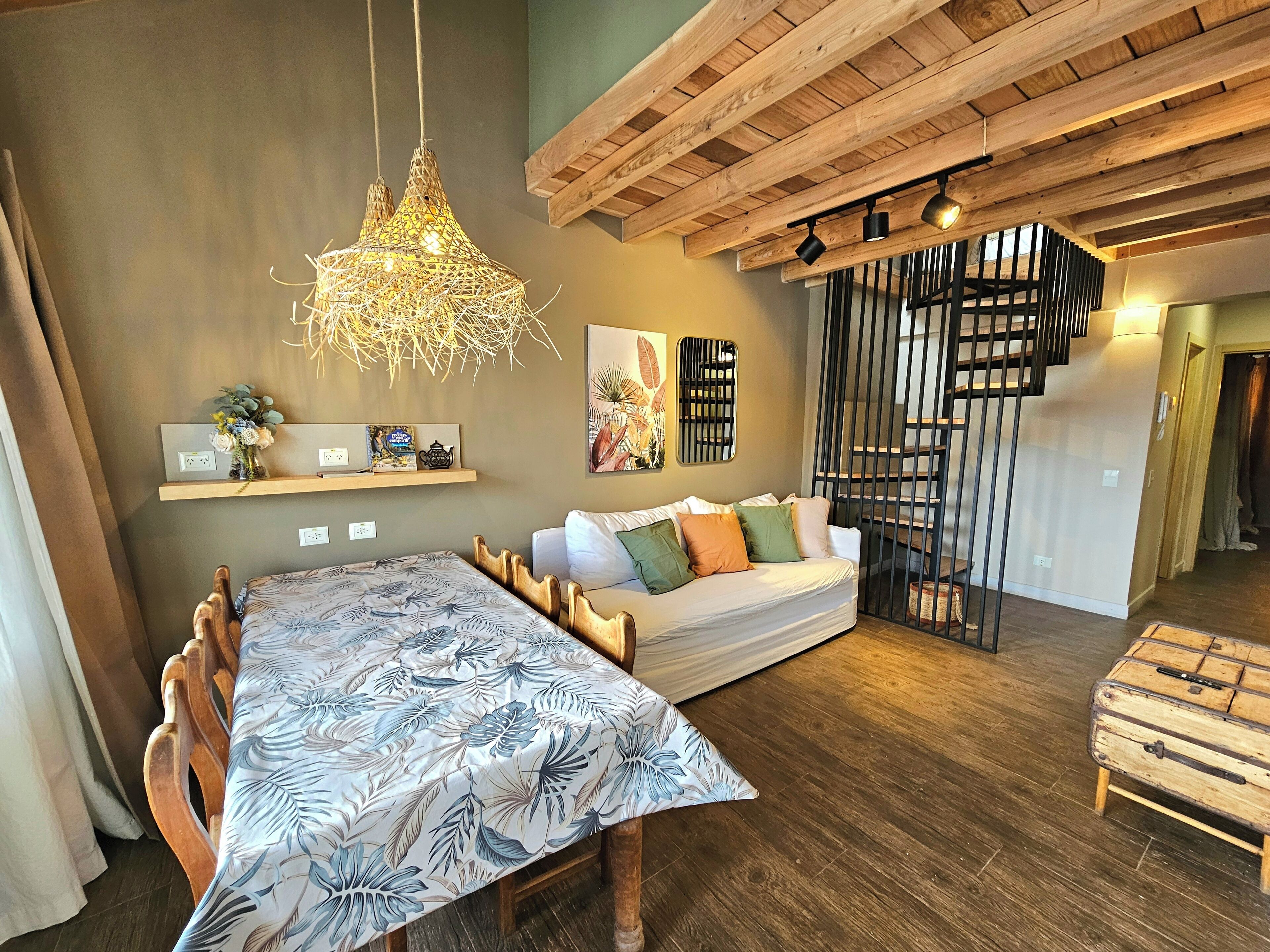 A modern living room with a wooden ceiling, a dining table with a floral tablecloth, a sofa with colorful cushions, and a staircase with a railing.