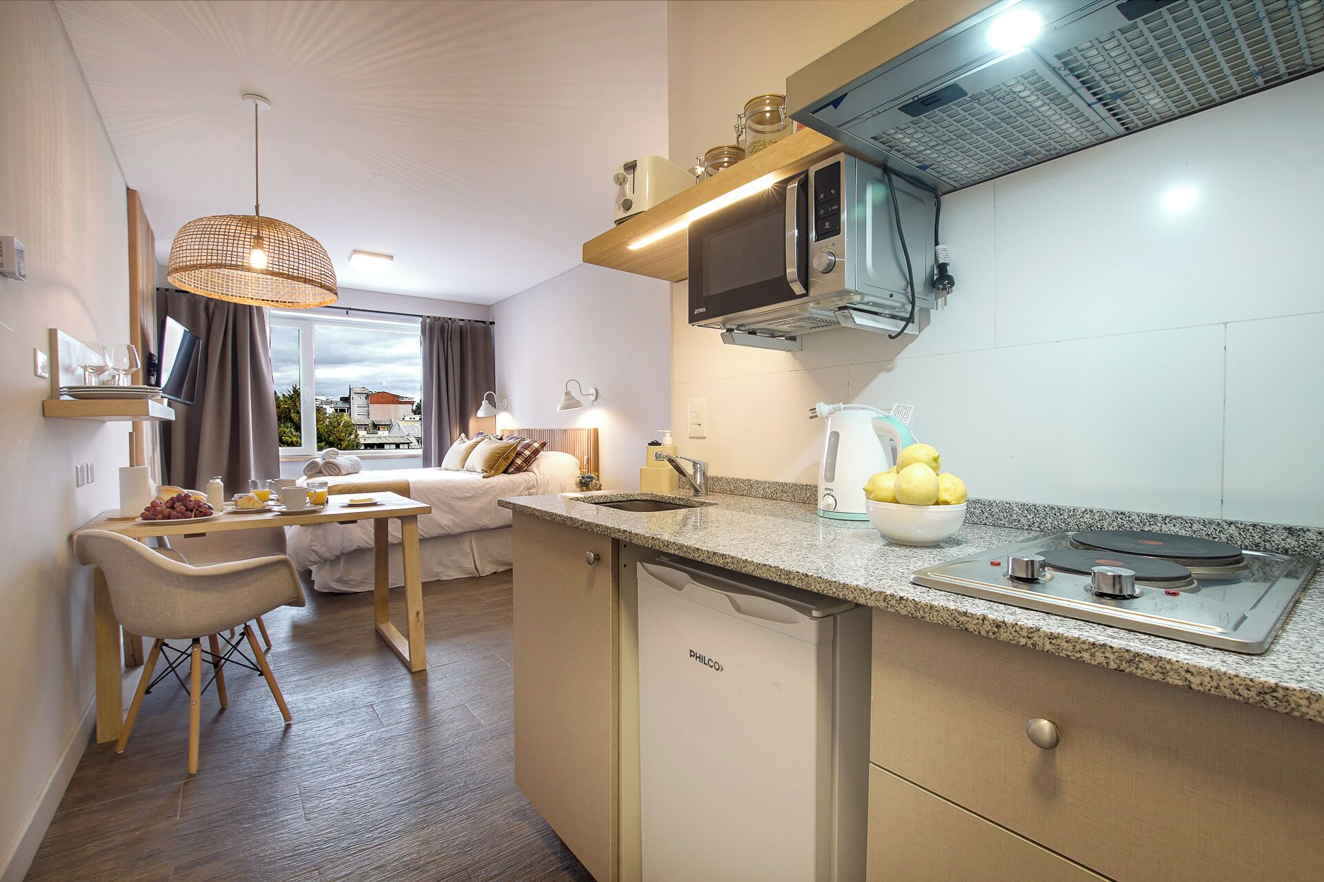 A compact kitchen with a built-in dishwasher, a microwave, and a kettle. There is a dining area with a table and chairs, and a bed with a view of the city.