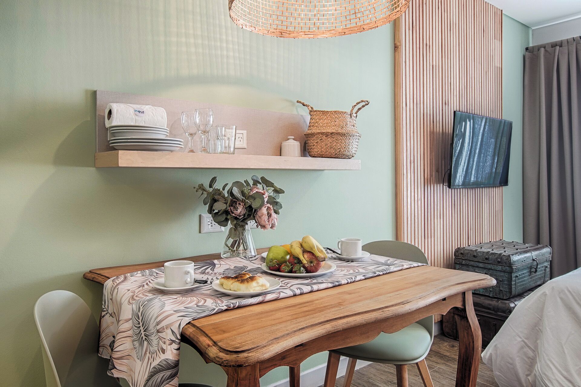 Un comedor con una mesa de madera, sillas, un florero con flores y un televisor montado en la pared.
