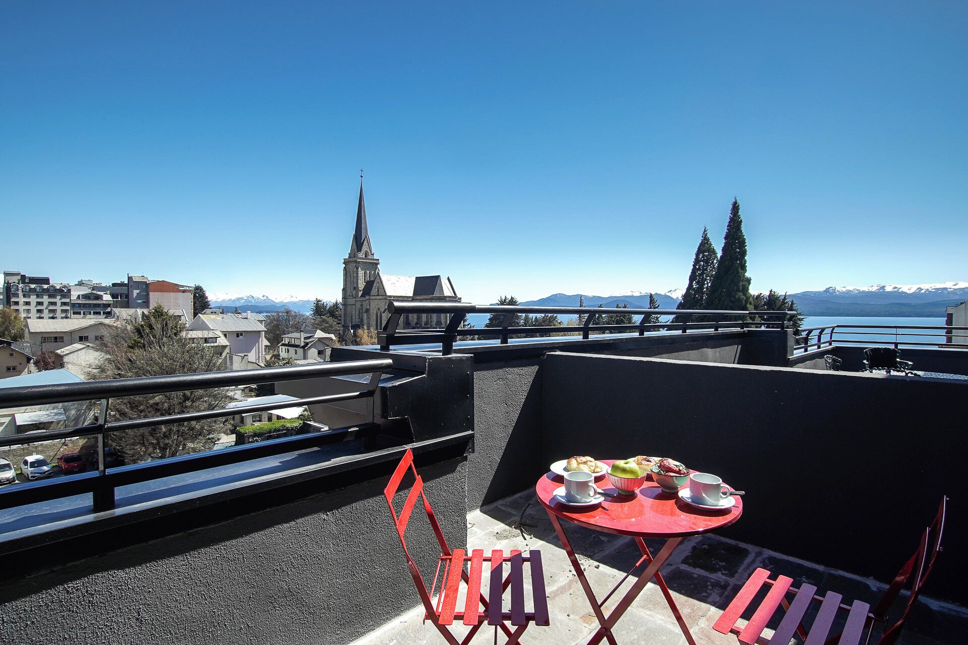 Una terraza en la azotea con una mesa lista para dos personas, con vistas a un paisaje urbano y un lago con montañas al fondo.