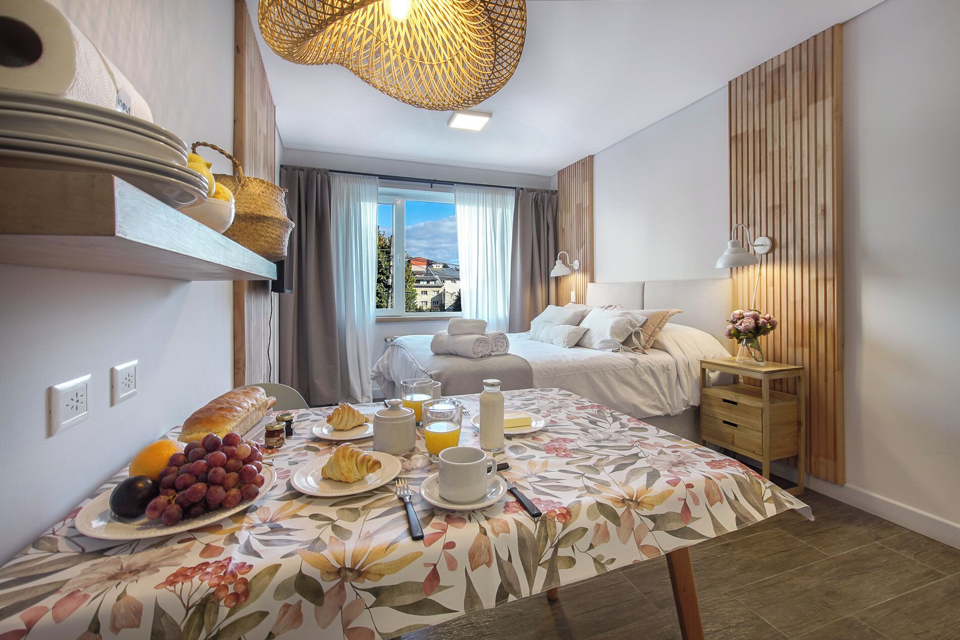 Habitación de hotel con una mesa de comedor lista para el desayuno, una cama con ropa blanca, cabecero de madera y vistas a edificios a través de la ventana.