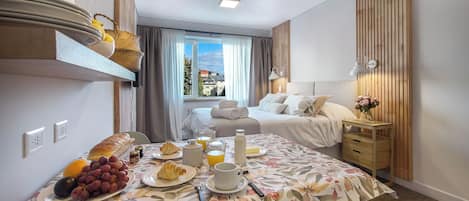 Habitación de hotel con una mesa de comedor lista para el desayuno, una cama con ropa blanca, cabecero de madera y vistas a edificios a través de la ventana.