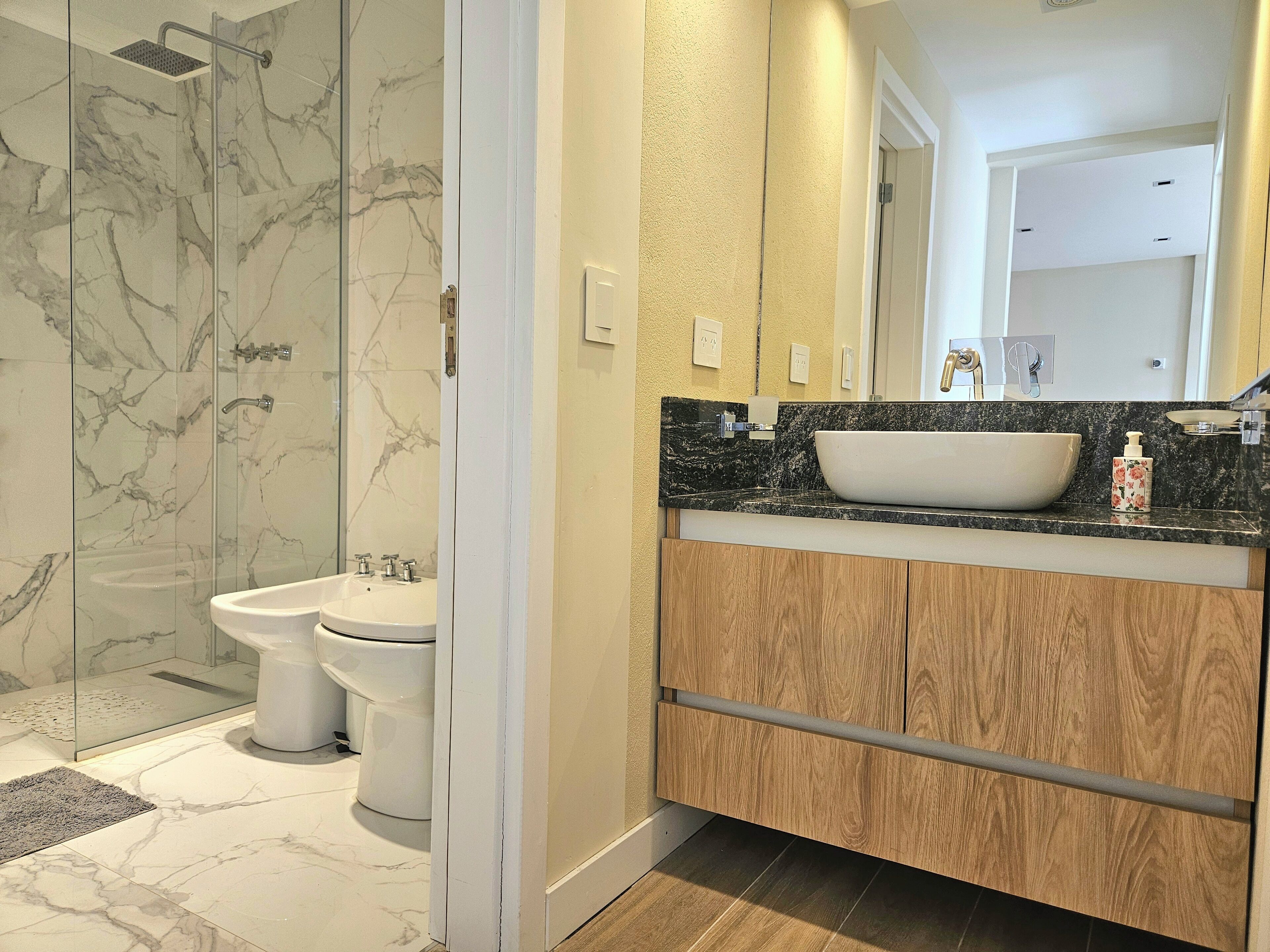 A bathroom with a glass shower enclosure, a white sink, and wooden cabinets.