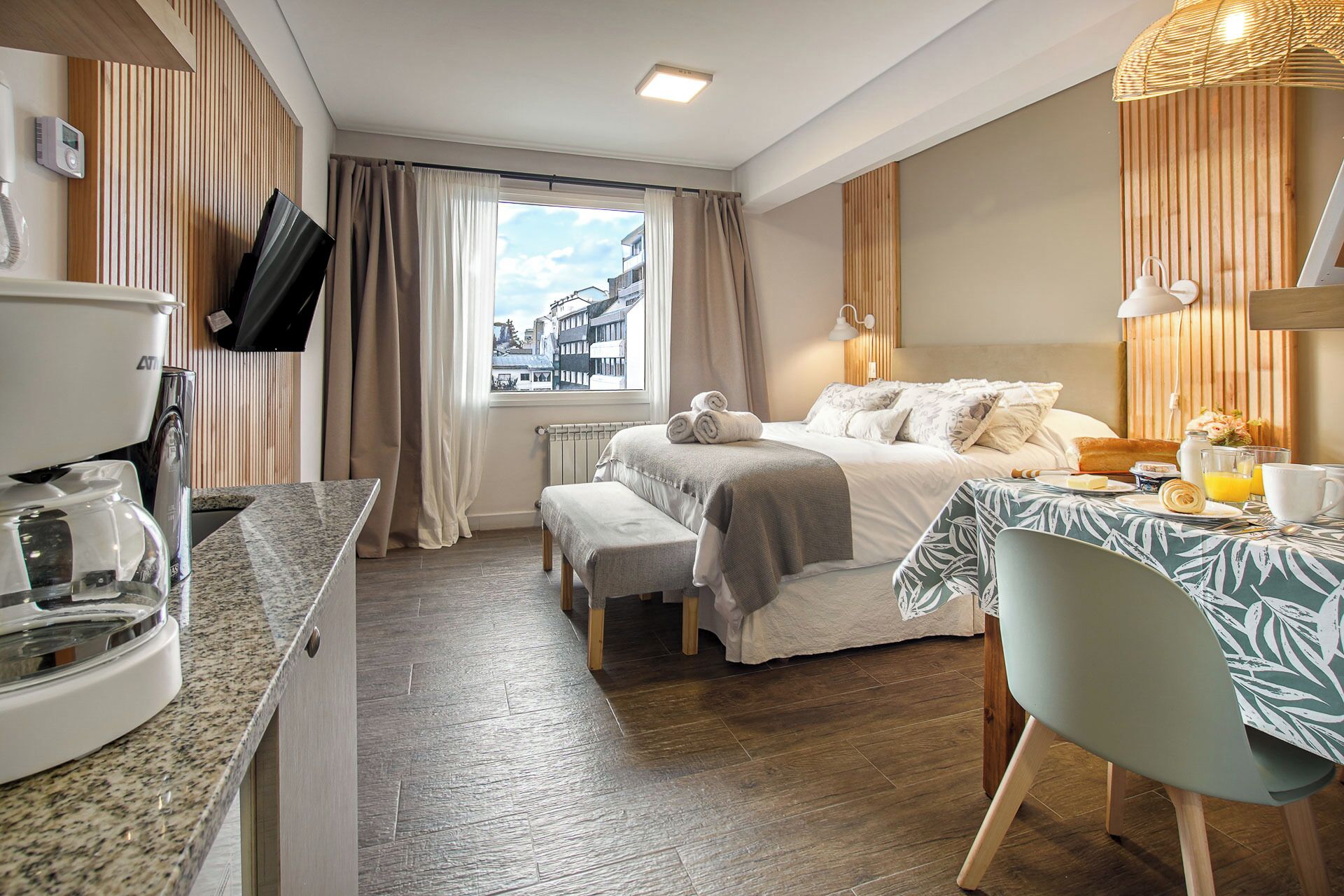 Habitación de hotel con cama, una mesa de comedor con desayuno, cafetera, televisor y ventana con cortinas.