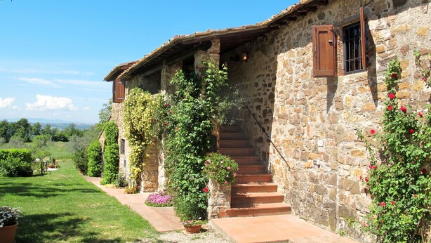 A stone building with a rustic facade, a set of stairs leading to a wooden door, and a garden with various plants and flowers.