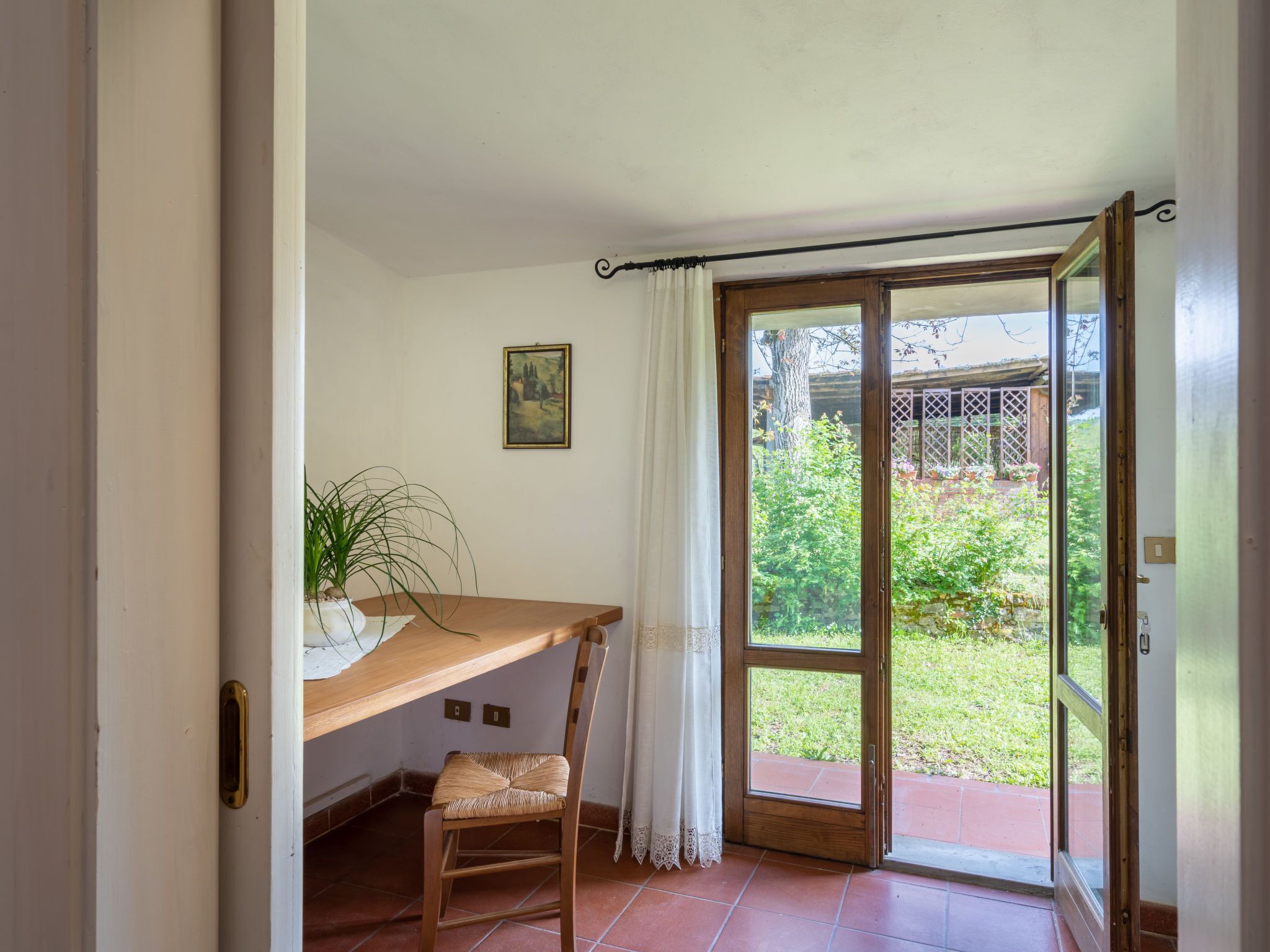 Een kamer met een houten bureau, een stoel, een potplant, een glazen schuifdeur die uitkomt op een tuin en een lijst met een afbeelding aan de muur.