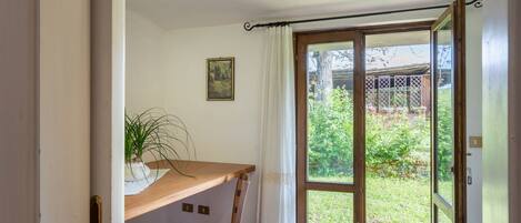 Een kamer met een houten bureau, een stoel, een potplant, een glazen schuifdeur die uitkomt op een tuin en een lijst met een afbeelding aan de muur.